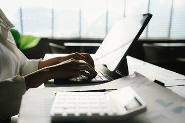 A person working with a calculator and financial documents on a desk, analyzing data from charts displayed on a laptop in a bright office environment.