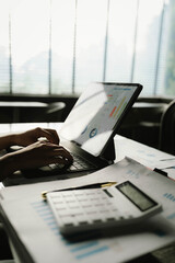 A person working with a calculator and financial documents on a desk, analyzing data from charts displayed on a laptop in a bright office environment.