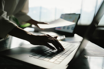 A person working with a calculator and financial documents on a desk, analyzing data from charts displayed on a laptop in a bright office environment.