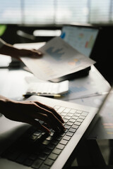 A person working with a calculator and financial documents on a desk, analyzing data from charts displayed on a laptop in a bright office environment.