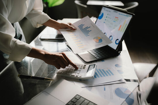 A person working with a calculator and financial documents on a desk, analyzing data from charts displayed on a laptop in a bright office environment.