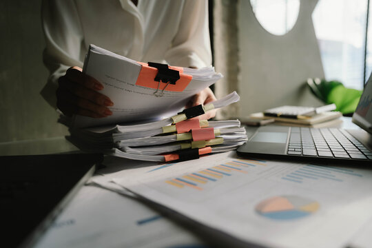 Close-up of businesswoman working at her desk with laptop and stack of documents, focusing on paperwork, reports and financial analysis in a busy office environment.