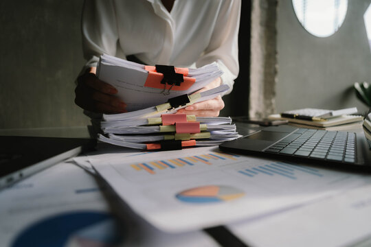 Close-up of businesswoman working at her desk with laptop and stack of documents, focusing on paperwork, reports and financial analysis in a busy office environment. - Powered by Adobe