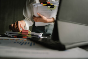 Close-up of businesswoman working at her desk with laptop and stack of documents, focusing on paperwork, reports and financial analysis in a busy office environment.
