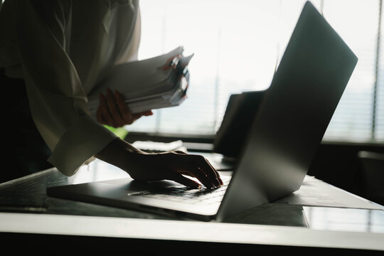 Close-up of businesswoman working at her desk with laptop and stack of documents, focusing on paperwork, reports and financial analysis in a busy office environment.