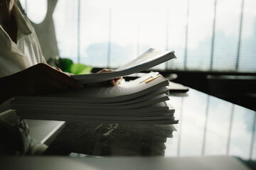 Close-up of businesswoman working at her desk with laptop and stack of documents, focusing on paperwork, reports and financial analysis in a busy office environment.