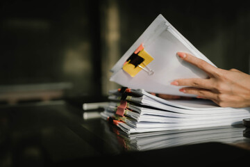 Close-up of businesswoman working at her desk with laptop and stack of documents, focusing on paperwork, reports and financial analysis in a busy office environment.
