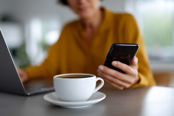 A realistic modern lifestyle photo in horizontal orientation (3:2 aspect ratio) showing a person working remotely, using a smartphone with one hand while sitting in front of a lapt
