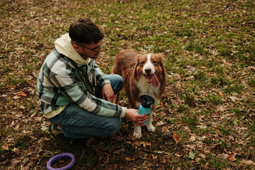 Man kneeling beside Australian Shepherd offering water from portable bottle after walk. Concept of care, friendship, and activity