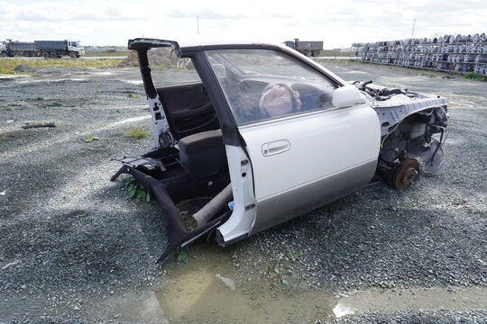 The cut-off side of the car at the car disassembly site. A wheel hub without a wheel, gravel and puddles, racks with wheels in the background.