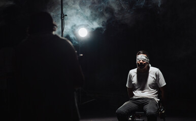 Man blindfolded and tied up being filmed in a dark room