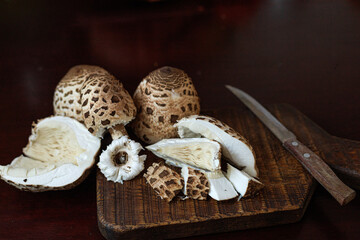 Macrolepiota procera, the parasol mushrooms, whole and sliced on a dark wooden board. Warm earthy tones, contrast of smooth white flesh and patterned brown caps. Concept of natural texture. © Natallia