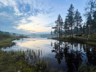 Morning fog lifting over a serene lake with pine trees and sunrise colors reflected in a peaceful natural landscape
