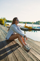 Young woman enjoying the serene lakeside atmosphere while seated on the wooden dock during a sunny...