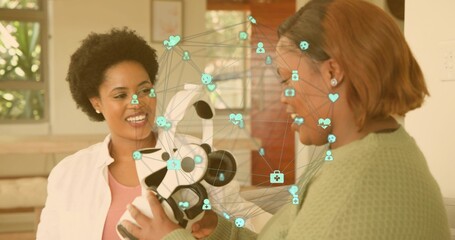 Holding white-black robot, two women in green sweater examining teal AR overlay in living room