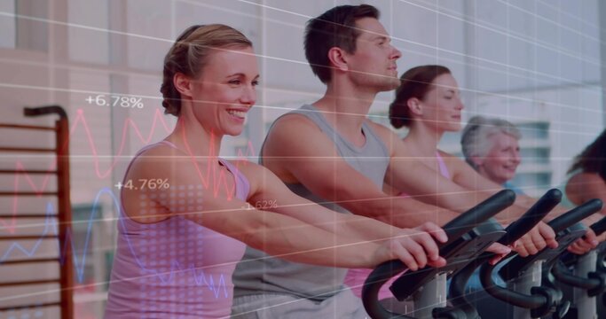 Pedaling group in athletic wear on exercise bikes at gym with stall bars, showing fitness metrics