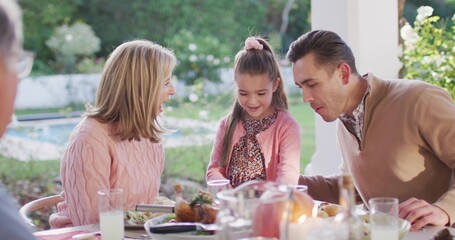 Standing girl in pink cardigan and floral dress leaning over covered patio table, adults reacting