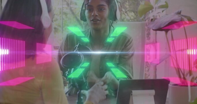 Recording host in denim-jacket and headset, speaking at table with cohost in yellow, mic, neon