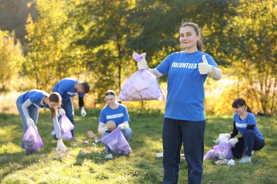 Young woman showing thumbs up and group of happy volunteers with bags collecting trash in park