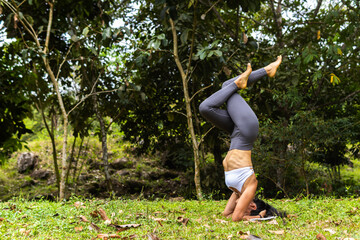 Athletic Latina woman performing a variation of an inverted yoga pose (Sirsasana) with crossed legs. The scene emphasizes core strength, balance, and outdoor wellness.