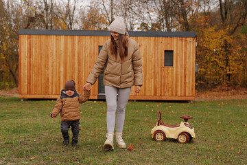 A mother holding her toddler son's hand walks in a grassy field, A ride-on toy car rests nearby, A...