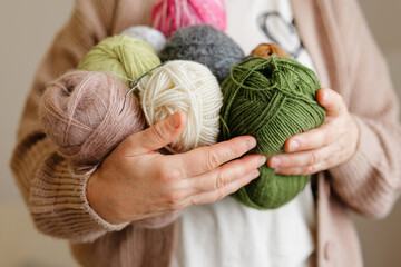 A close-up shot of a person's hands holding a stack of yarn balls in various neutral and earthy colors, including pink, green, white, and gray, ready for crafting.