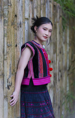 Fototapeta premium Young woman in traditional ethnic dress with colorful embroidery and pom poms leans against rustic wooden wall, expressing calm confidence