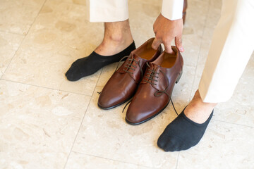 A man is preparing for a formal occasion by putting on his brown dress shoes. He wears black socks and stands on light-colored marble flooring in a well-lit space, exuding elegance.