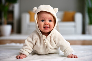Cute Caucasian baby with blue eyes looks playfully surprised while lying on her tummy in a cozy beige fleece jumpsuit with bear ears in a bright, modern home.