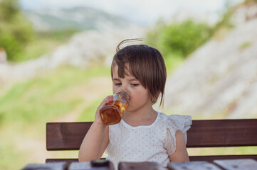A little girl is drinking juice while sitting on a wooden bench somewhere outdoors in nature	
