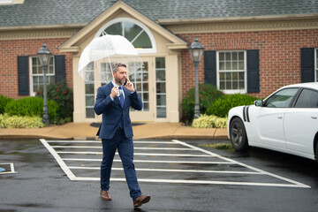 Businessman talks on the phone while standing in the rain. Man in suit with umbrella and makes a mobile phone call. Business man checks his smartphone on a rainy street. Call while rain drops fall.