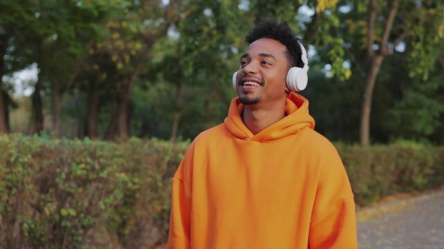 Side view young happy calm man he wear orange hoody listen to music in headphones dance walking rest relax in spring green city park go down sunshine alley outdoors on nature. Urban leisure concept