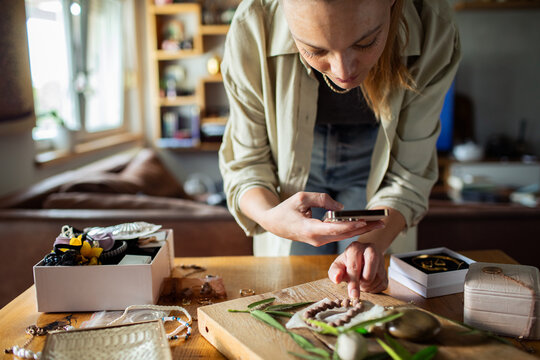 Young adult woman focused photographing jewelry with smartphone at home - Powered by Adobe