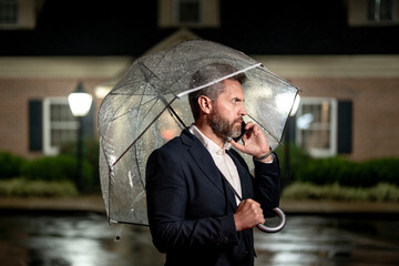 A businessman talks on his phone under an umbrella on a rainy night. Manager makes a business call in the rain. Man in suit speaking on a mobile phone in night street. Business man checks his schedule