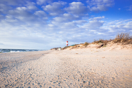 A tranquil coastal landscape featuring a sandy beach, driftwood, and lighthouse. Soft sunset light washes the scene in warm tones, with gentle waves and a blue sky.