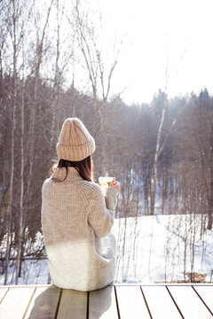 A woman wearing a cream knit sweater and matching beanie sits on a wooden deck in a snowy landscape, sipping a warm beverage and enjoying a quiet winter moment outdoors
