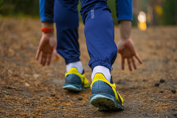 Runner's lower body in athletic shoes and tracksuit trousers, preparing for a sprint or jog on a natural path, hands touching the ground in a starting block position