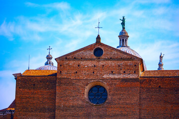 Facade of a historic brick building in Padua, featuring large circular window and several domes topped with crosses and statues. Blue sky highlight building historical and cultural significance