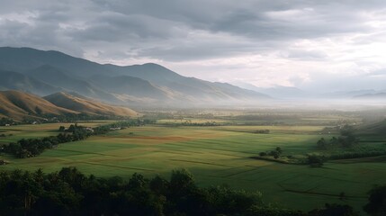 Obraz premium Misty valley landscape with rolling hills agricultural fields and distant mountains under atmospheric light