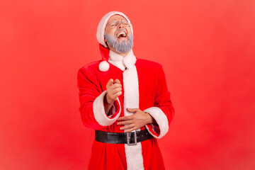 Happy funny elderly man with gray beard wearing santa claus costume pointing to camera with index finger, holding belly, laughing at you.