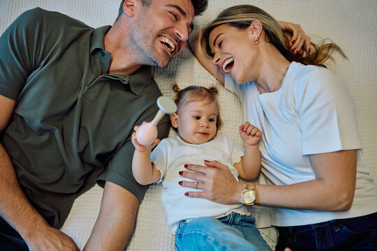 Happy family lying on bed, parents smiling and looking at their little baby girl holding a toy rattle
