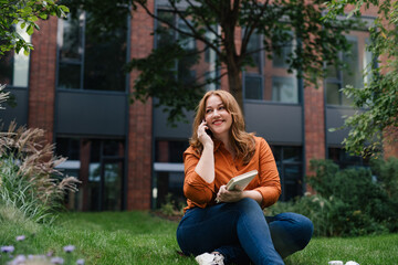 Confident woman rmaking phone call while reading book outdoors in park.