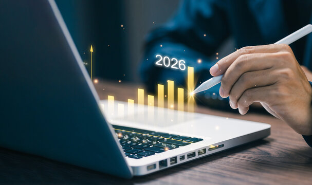 Business hand using stylus to draw upward yellow growth bar chart labeled 2026 on laptop. Concept of future financial growth, investment trend, and business success analytics.