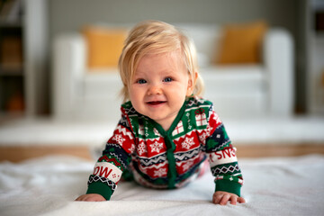 Cute Caucasian baby girl with blue eyes smiles widely while lying on her tummy in a cozy, festive Christmas jumpsuit in a bright, modern home