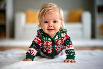 Cute Caucasian baby girl with bright blue eyes smiles widely while lying on her tummy in a cozy, festive Christmas holiday jumpsuit in a warm home setting.