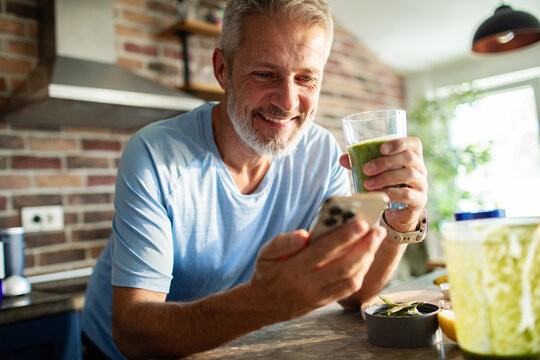 Mature man smiling while drinking smoothie and using smartphone in home kitchen - Powered by Adobe