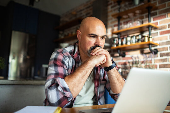Adult man looking thoughtful at laptop in home kitchen