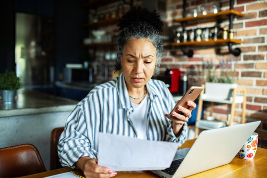 Mature woman worried while checking bill on phone at home kitchen