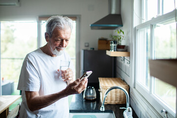 Senior man smiling at smartphone while drinking water in home kitchen