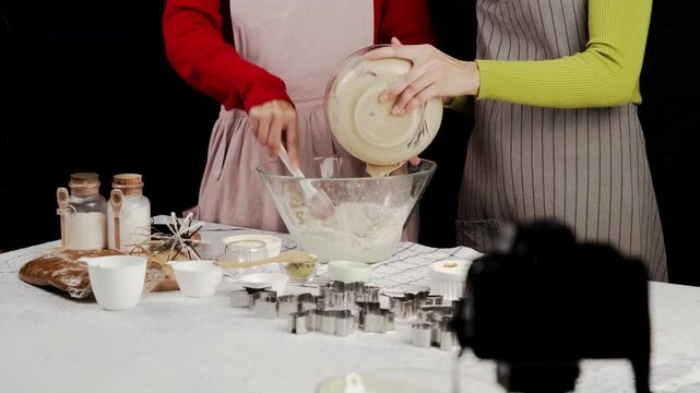 Two women in aprons are combining cookie batter and flour, preparing dough together on a kitchen counter filled with baking ingredients, cookie cutters, and tools